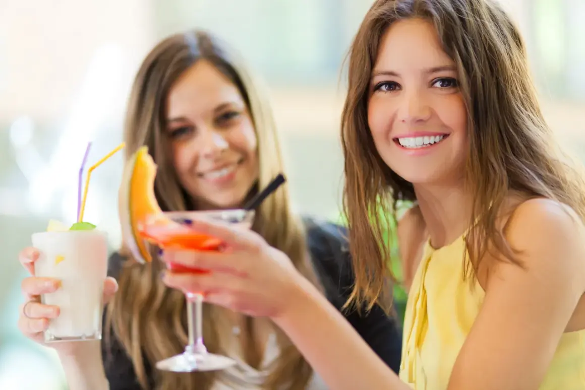 Dwie uśmiechnięte kobiety z drinkami. Jedna trzyma biały koktajl, druga pomarańczowy. Czy wódka tuczy?