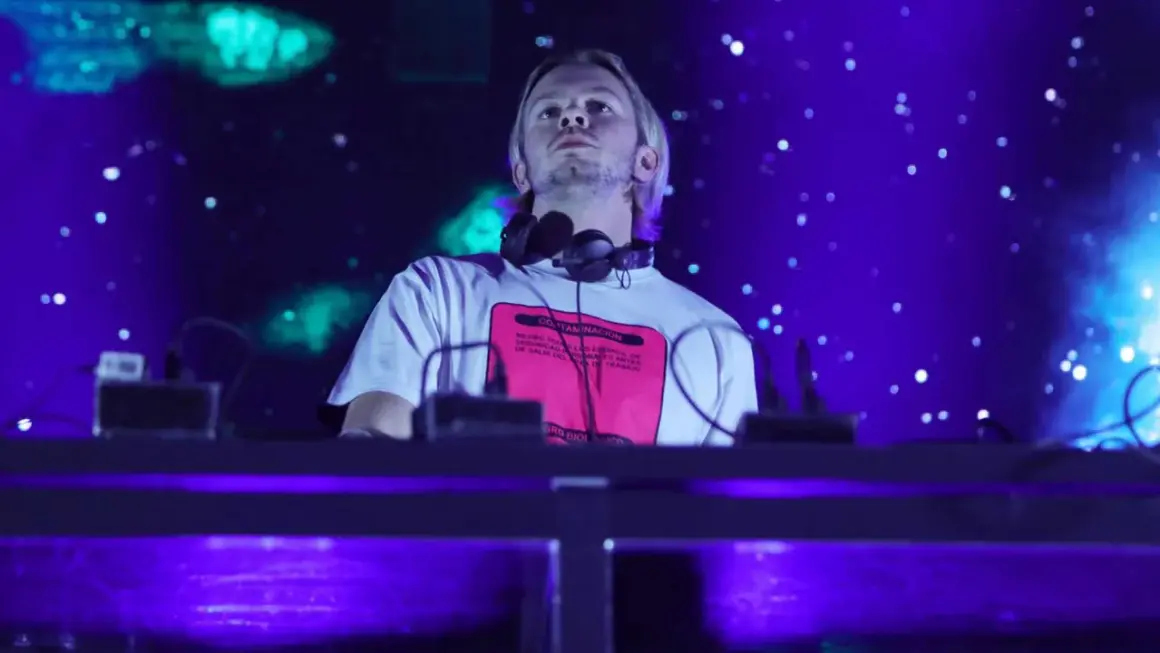 DJ na scenie, w słuchawkach na szyi, z napisem na koszulce. W tle niebieskie światła i efekty. To część lineupu Wisłoujście.