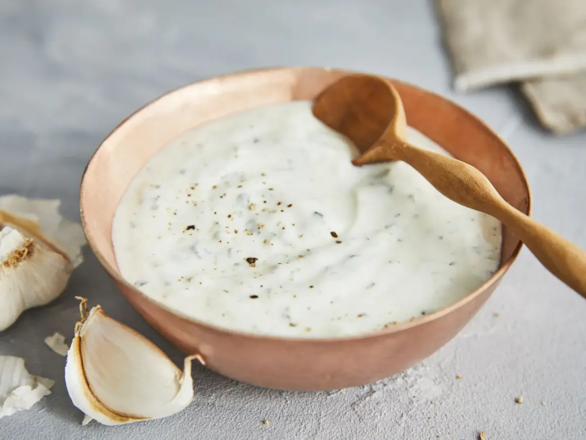 Ein cremiger Knoblauch Joghurt Dip in einer Kupferschale, garniert mit Pfeffer und Kräutern. Daneben liegen frische Knoblauchzehen.