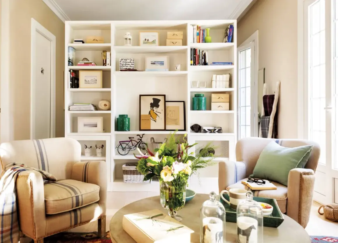 Acogedor salón con estanterías blancas llenas de libros y objetos decorativos, dos sillones de rayas y una mesa de centro con flores. Un rincón que evoca la calidez de las casas pequeñas.