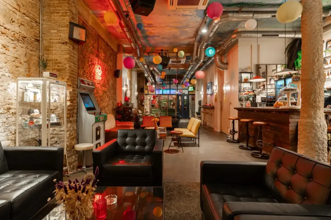 Interno di un social club a Barcellona con divani neri, un cabinato arcade, lanterne colorate e un bancone bar in legno.