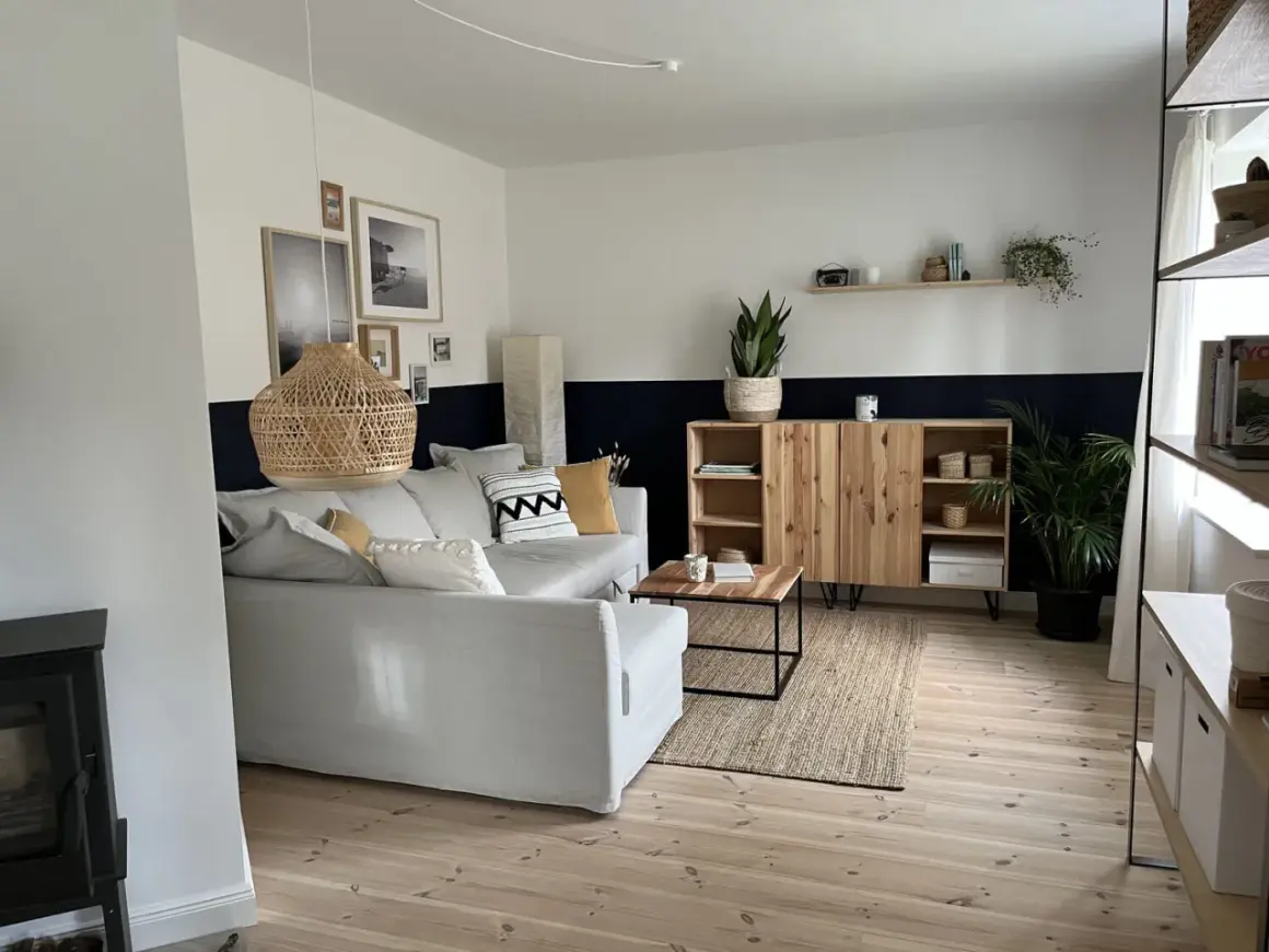 Gemütliches Wohnzimmer mit zweifarbig gestrichener Wand, die ein schönes Wandfarbe Muster bildet. Ein hellgraues Sofa, ein Holzschrank und ein Korb-Hängelampen runden den Look ab.