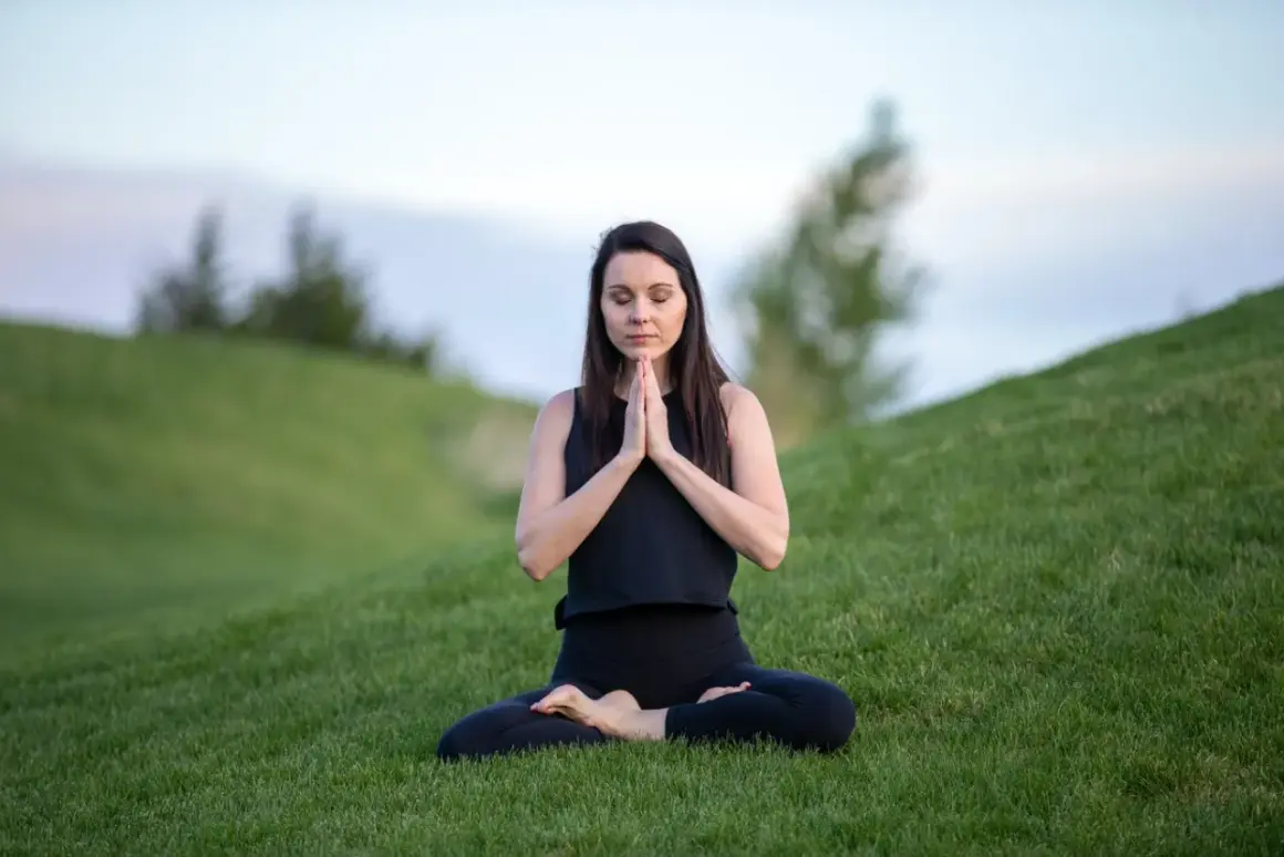 Mujer en posición de loto en la hierba, con las manos juntas en oración. Su rutina de yoga al aire libre es un momento de paz.