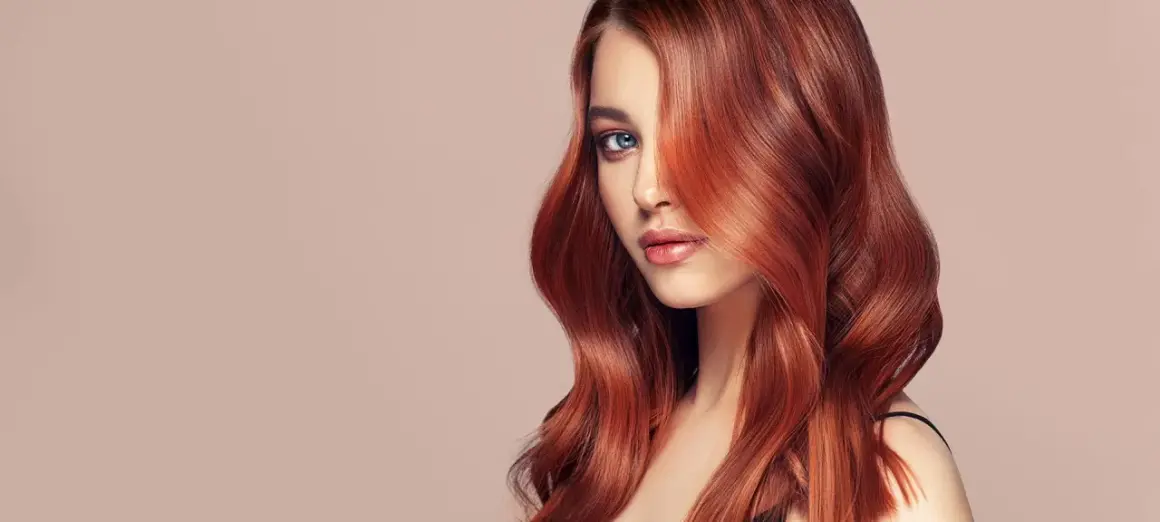 Ein Porträt einer Frau mit voluminösen, dunkelroten Haaren, die ihr Gesicht umspielen. Ihre blauen Augen blicken nachdenklich in die Ferne.