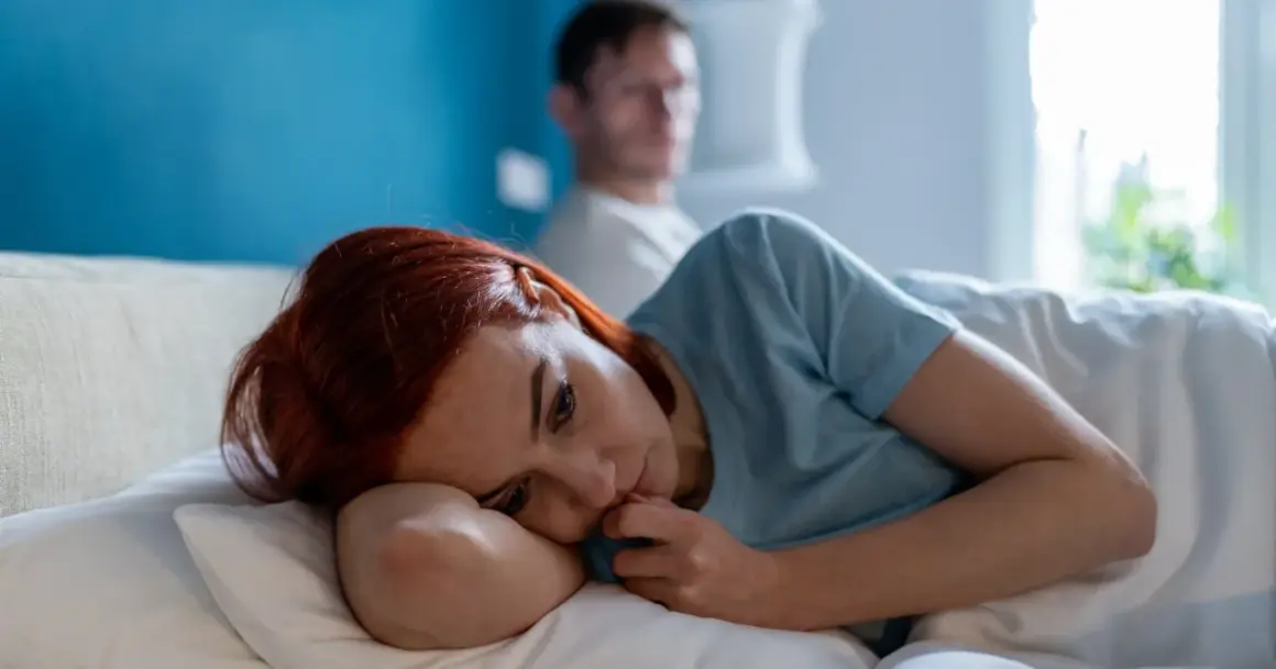Smutna kobieta leży w łóżku, czując się jak ofiara w związku. Jej partner siedzi za nią, odwrócony plecami.