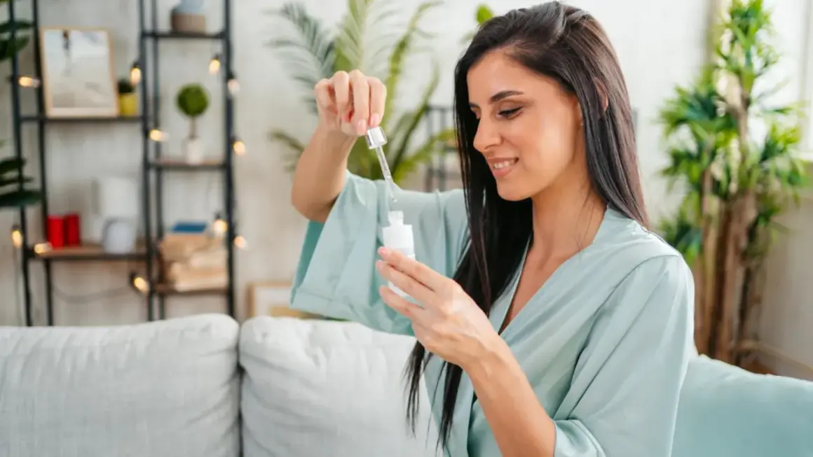 Frau mit dunklen Haaren in einem hellblauen Morgenmantel, die einen Tropfen eines Vitamin C Serums aus einer Pipette auf eine Flasche fallen lässt.