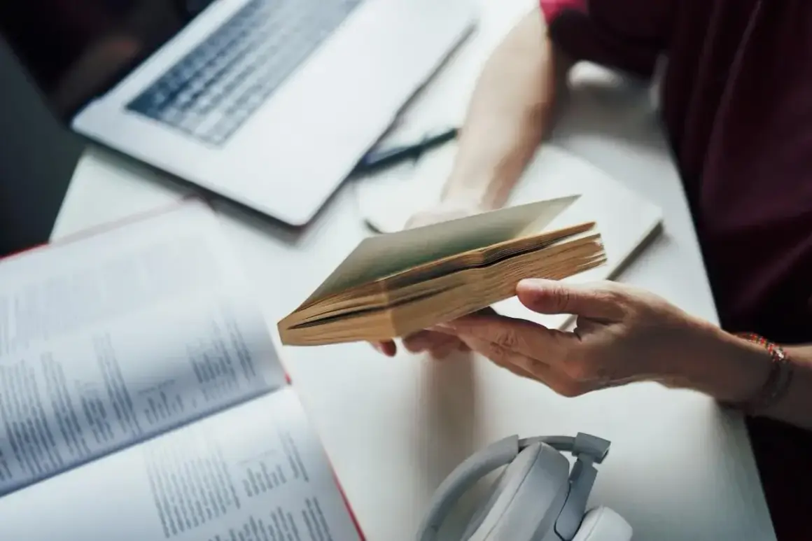 Ręce trzymają otwartą książkę, obok laptop i słuchawki. To dobry przykład, jak tworzyć bibliografię.