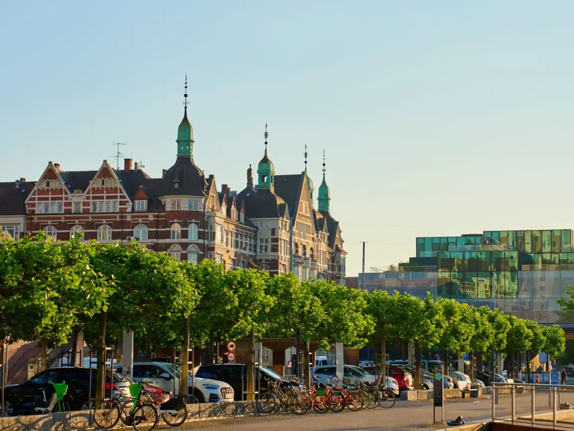 Kopenhaga ceny: zabytkowe budynki i nowoczesna architektura nad wodą, z rzędem drzew i zaparkowanymi samochodami.