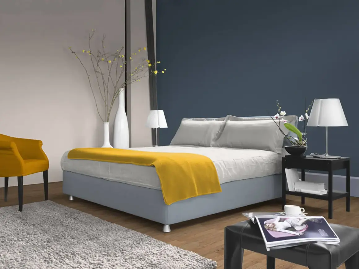 Ein Schlafzimmer mit einem Bett, das mit einer gelben Decke und Kissen in verschiedenen **blau nuancen** dekoriert ist. Ein gelber Sessel und ein Teppich ergänzen den Raum.