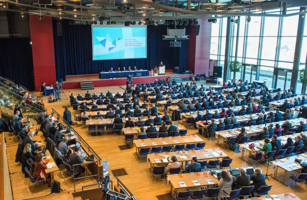 Große Versammlung mit vielen Zuhörern, die auf eine Bühne blicken. Die Bedeutung einer Synode wird hier durch die Präsentation und die Anwesenheit vieler Menschen verdeutlicht.