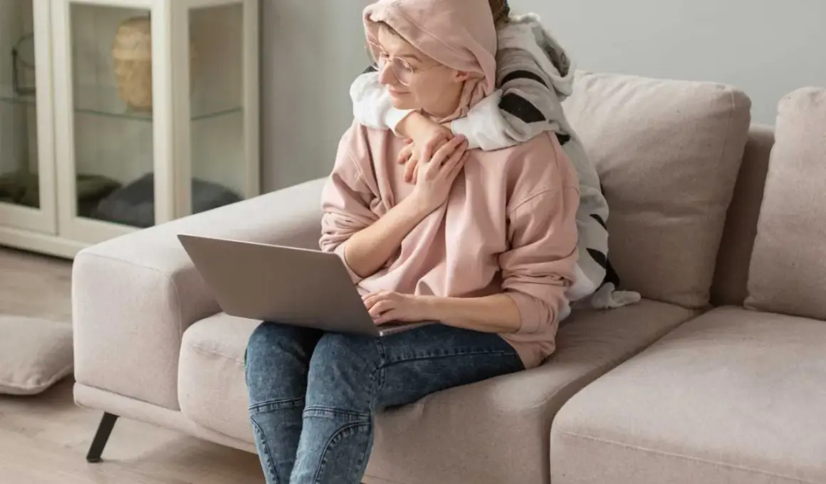 Kobieta z dzieckiem na kolanach pracuje przy laptopie. Dziecko ją przytula, szukając wsparcia, jak u psychoonkologa.