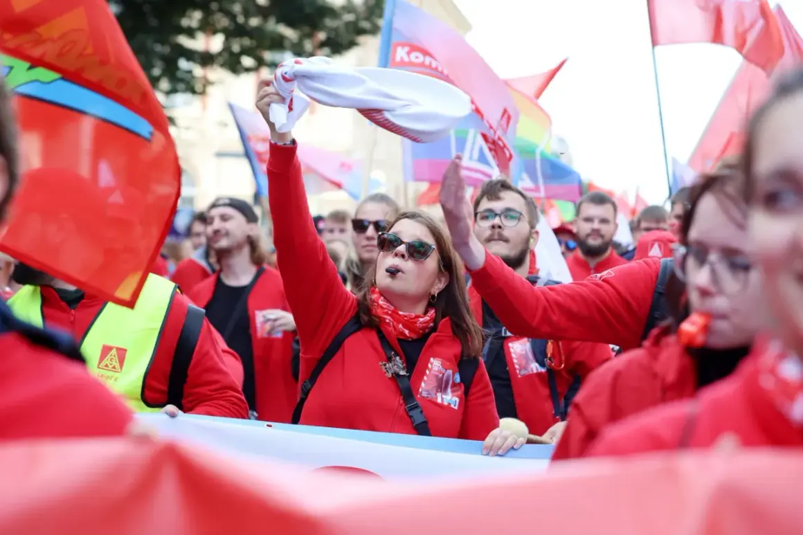 Frau mit Pfeife und weißem Tuch winkt, umgeben von roten Fahnen und Menschen bei einer Demonstration. Sie ist die Vorsitzende der IG Metall.