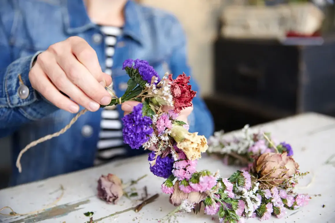 Hände binden bunte Trockenblumen zu einem Kranz. Ein schönes Projekt, um Trockenblumen selbst zu machen.