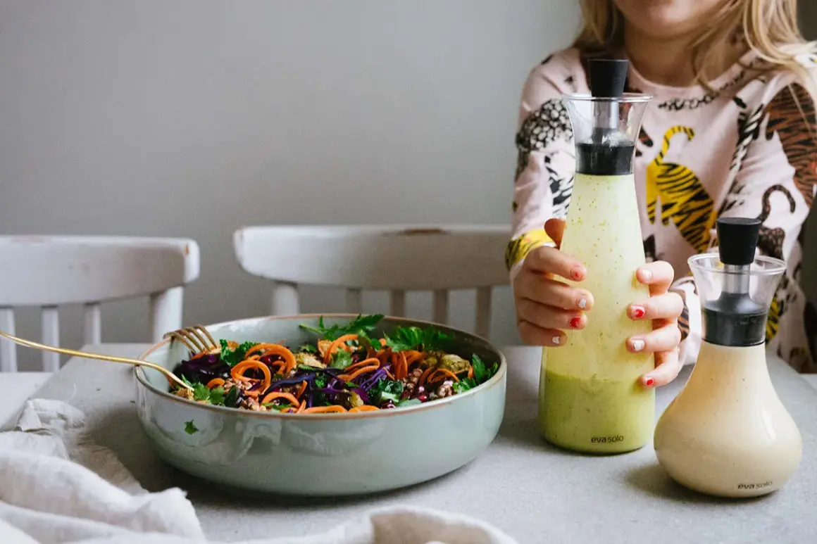En färgstark sallad med morötter och rödkål i en skål, bredvid två flaskor med dressing till sallad. Ett barn håller i den ena flaskan.