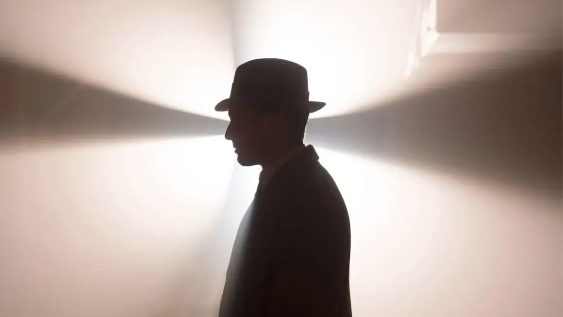 Ein Mann im Hut und Anzug, im Profil, vor einem hellen, strahlenden Hintergrund. Ein mysteriöser Blick, der an Babylon Berlin Staffel 5 erinnert.
