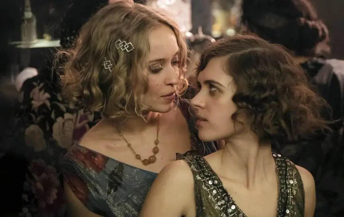 Zwei Frauen im Stil der 1920er Jahre, eine mit blonden Locken und Schmuck, die andere mit dunklen Locken, blicken sich intensiv an. Ein Moment aus Babylon Berlin Staffel 5.