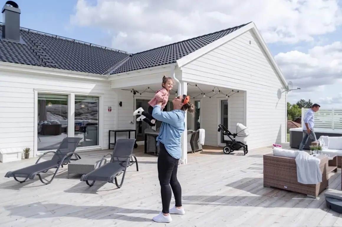 En kvinna lyfter sitt barn på en altan vid ett modernt hus, en idyllisk semesterbild från ett resort i Sverige.