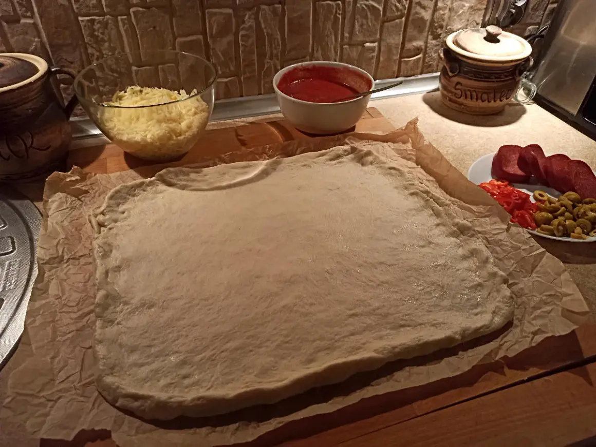 Gotowe ciasto na pizzę czeka na dodatki: ser, sos, salami, papryka i oliwki.