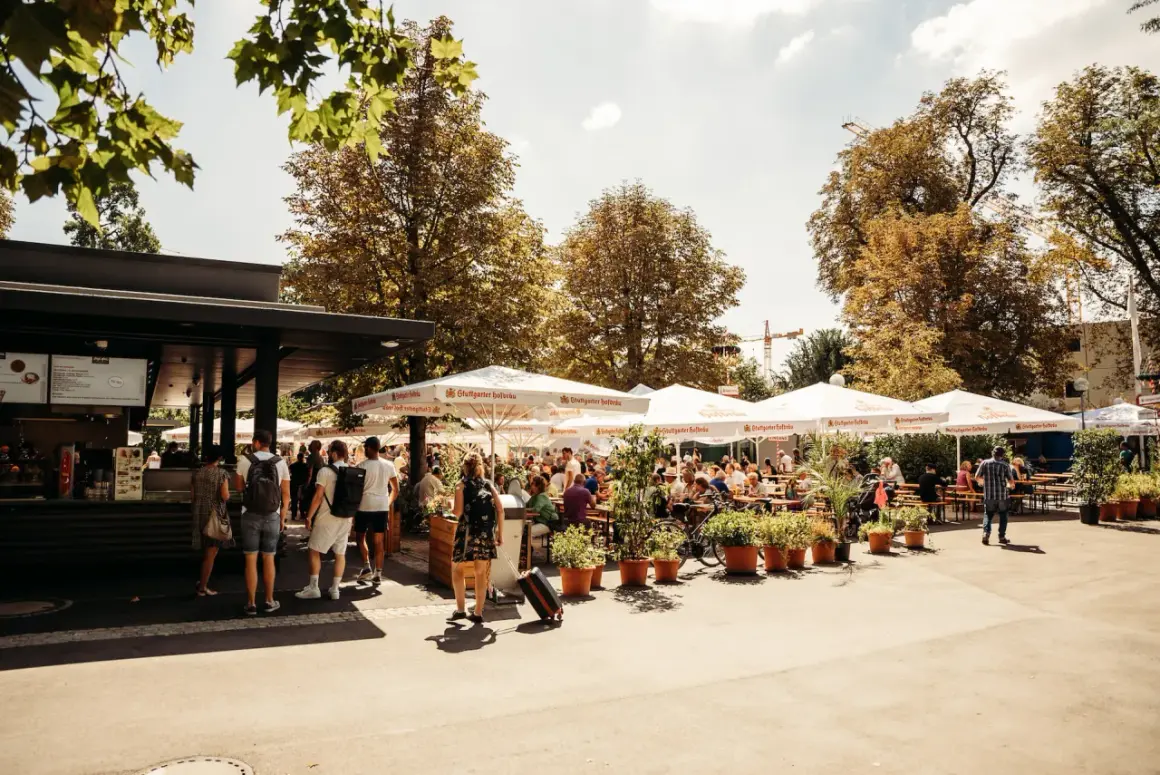 Sonniger Biergarten im Schlossgarten mit vielen Gästen unter schattenspendenden Schirmen.