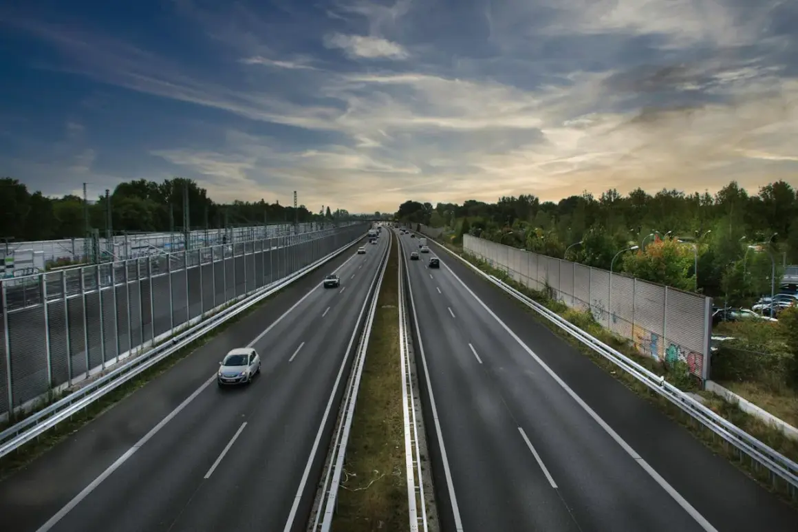 Prowadzenie pojazdu po szerokiej autostradzie, z samochodami w oddali i drzewami po bokach.