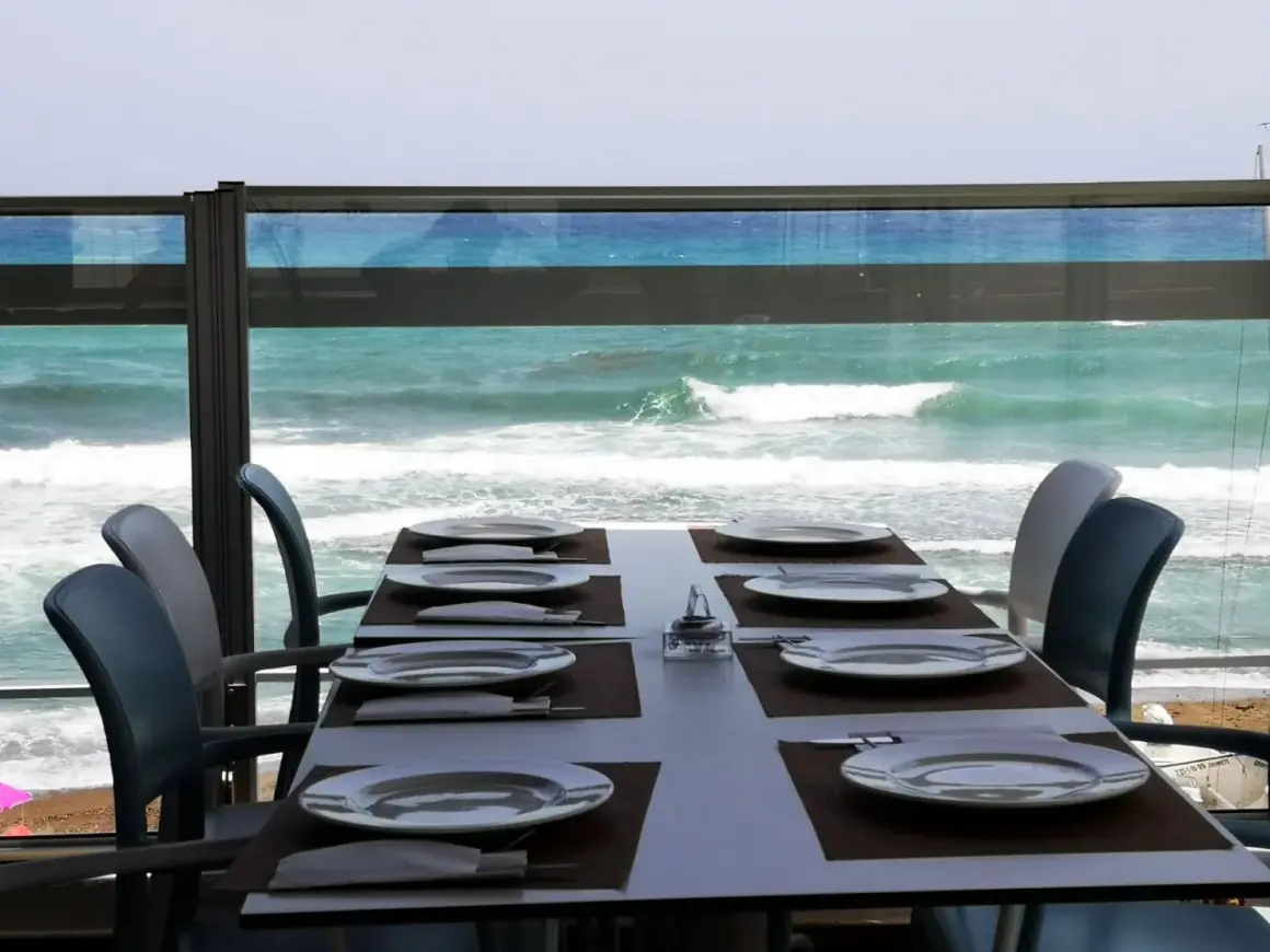 Mesa puesta en el chiringuito la sal, con platos y cubiertos, frente a un mar turquesa y olas rompiendo.