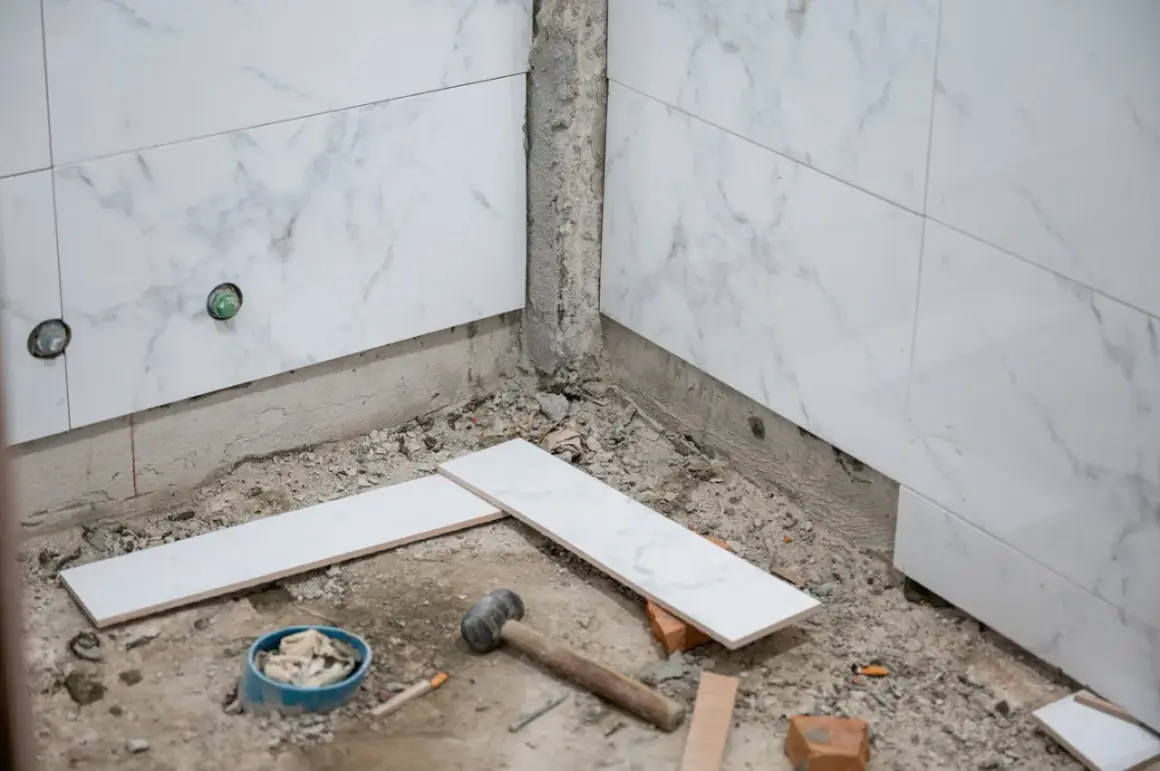 Début des travaux pour carreler une salle de bain : des carreaux de marbre blanc sont posés sur le sol, près d'un marteau et d'un seau.