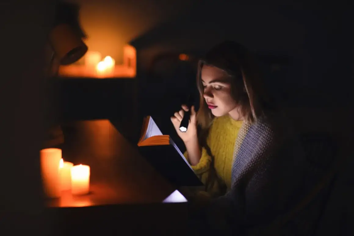 Dziewczyna czyta książkę przy świecach i latarce podczas blackoutu. Co to blackout? To nagłe przerwy w dostawie prądu.