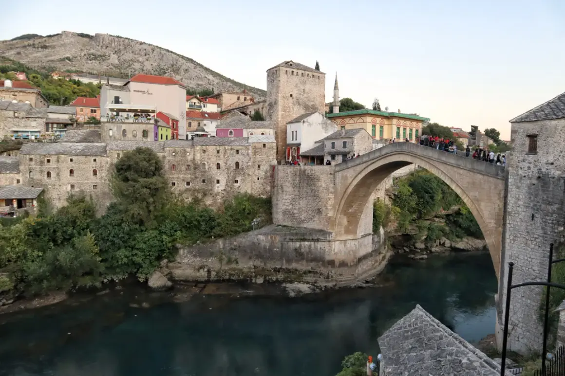 Kamienny, łukowy Mostar most nad turkusową rzeką, otoczony zabytkową zabudową i zielenią.
