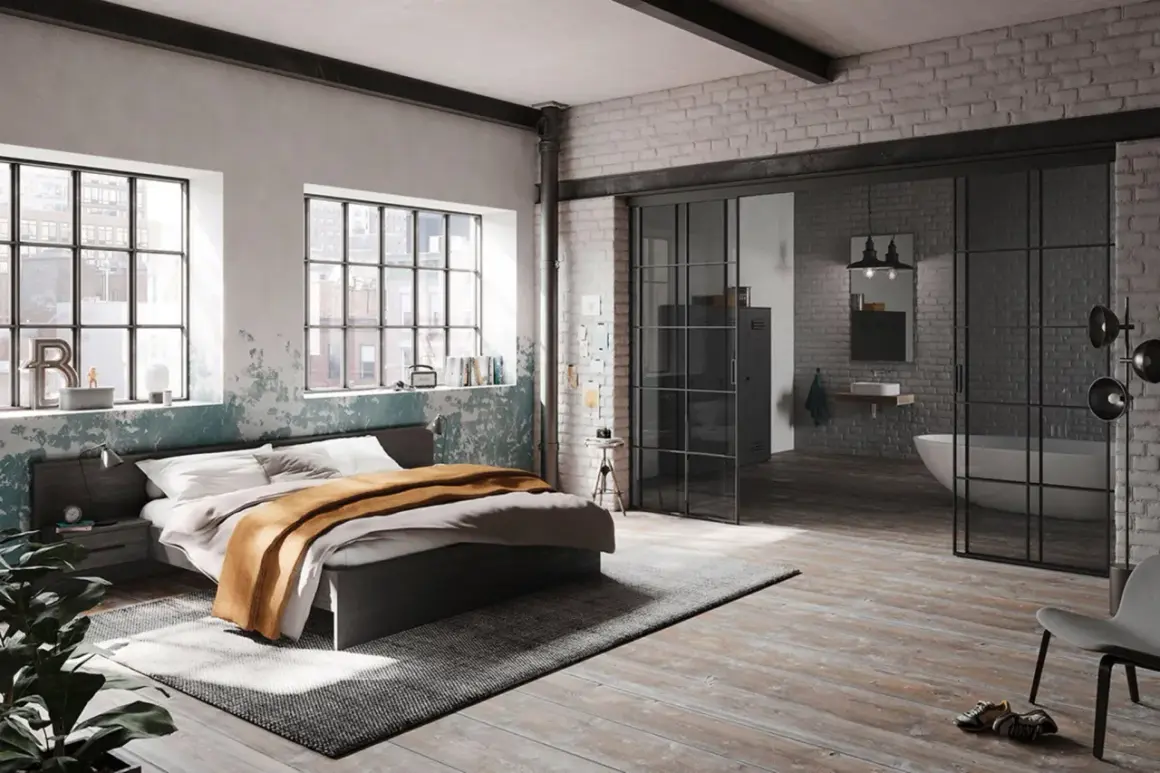 Ein gemütliches **Bett unter Fenster** mit industriellem Charme. Daneben ein offener Badbereich mit Wanne.