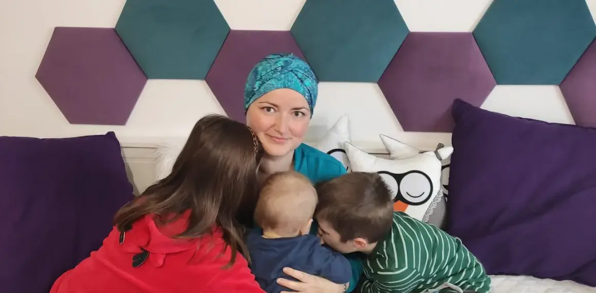 Mama w chustce i trójka dzieci. Najstarszy syn całuje maluszka, córka przytula się do mamy. W tle heksagony i poduszki. BIRADS 4a.