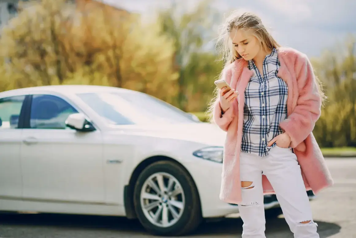 Młoda kobieta w różowym futrze i podartych dżinsach sprawdza telefon obok białego samochodu, być może po **szkodzie parkingowej**.