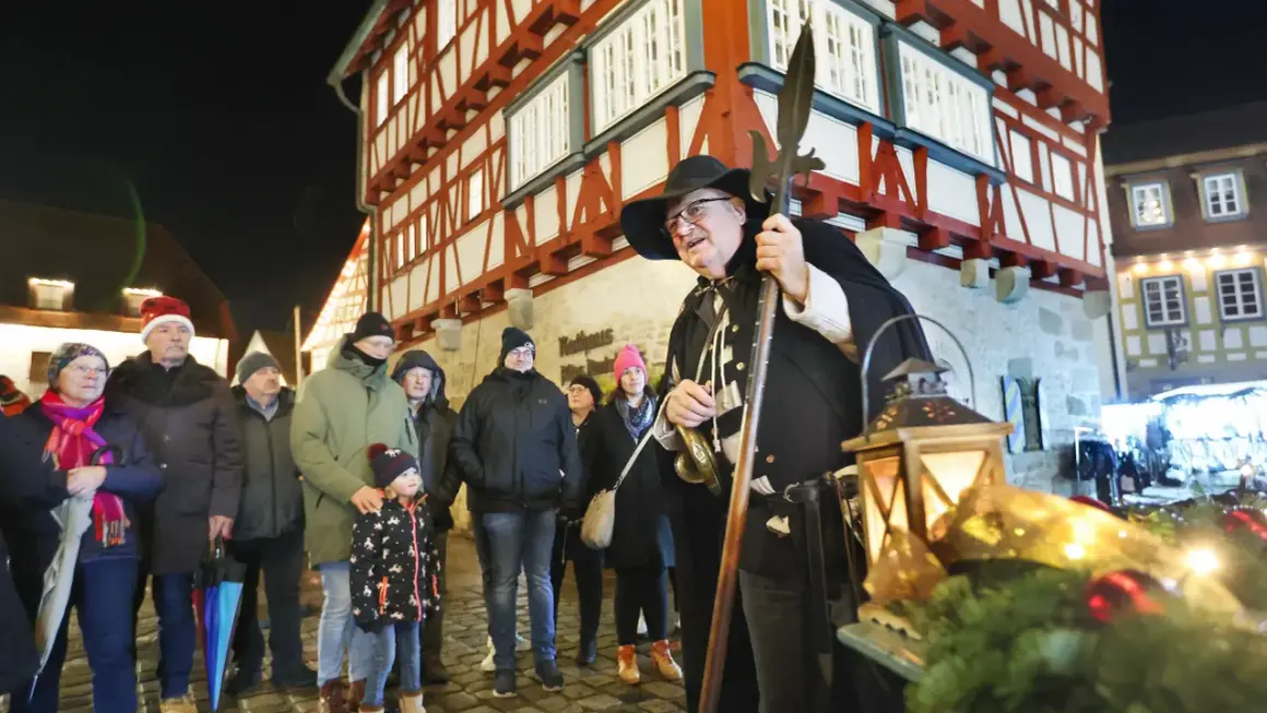 Ein Nachtwächter mit Hellebarde und Laternen auf dem Vellberg Weihnachtsmarkt. Besucher in Weihnachtsmützen und warmen Jacken stehen davor.