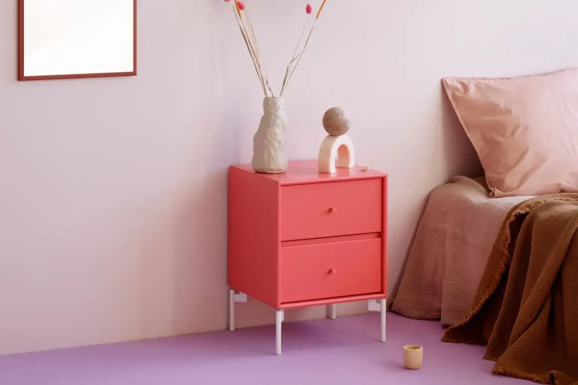 Ein Nachttisch in leuchtender **pink Farbe** steht neben einem Bett mit rosa Kissen. Dekorative Objekte und eine Vase mit Trockenblumen zieren die Oberfläche.
