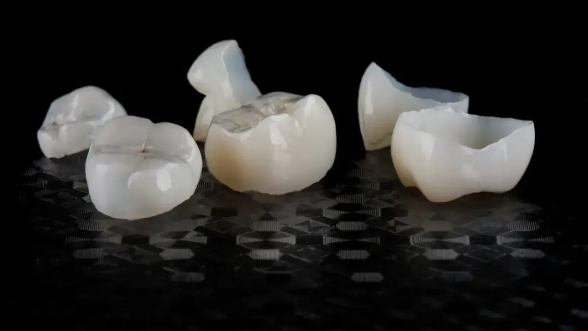 Kilka białych, ceramicznych koron dentystycznych leży na czarnym, błyszczącym tle.