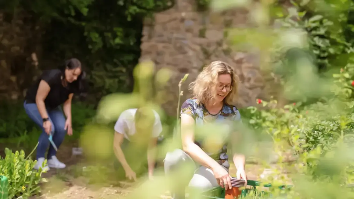 Drei Menschen im Garten, die pflanzen. Die Frau vorne lacht, während sie Töpfe sortiert. Hier im Grünen, bei der Arbeit, fühlt sie: warum lebe ich.