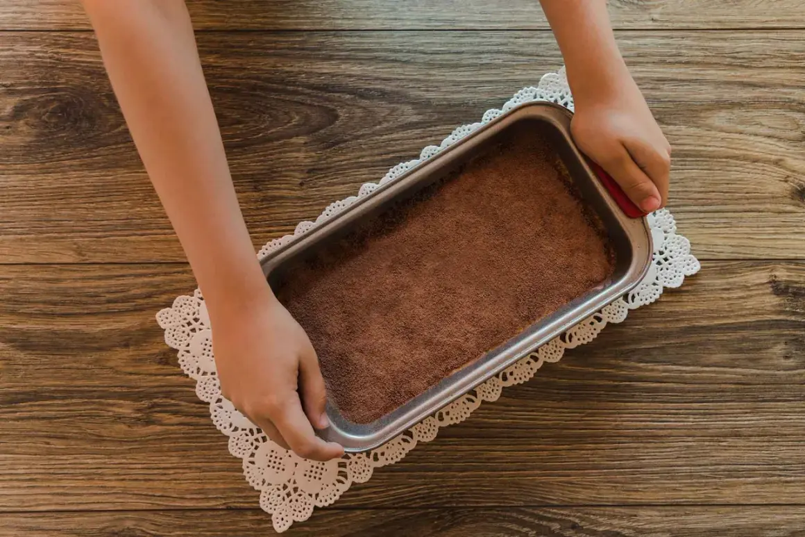 Mani che preparano un tiramisù senza caffè, con cacao in polvere in una teglia su un centrino di pizzo.