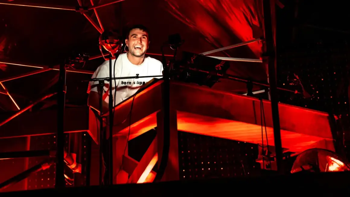 Il **best techno dj** ride dietro la console, immerso in luci rosse intense.