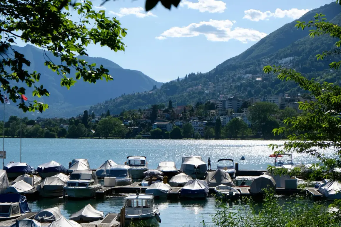 Båtar täckta med presenningar vid en brygga vid Lago Maggiore. Berg och en stad syns i bakgrunden.