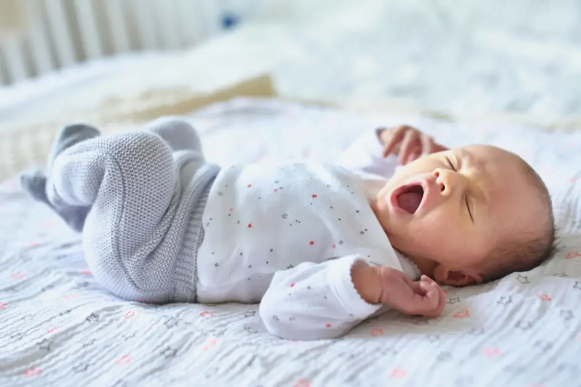Ein Baby gähnt auf einer Decke mit Sternen. Wann Babys alleine einschlafen, ist eine Frage, die viele Eltern beschäftigt.