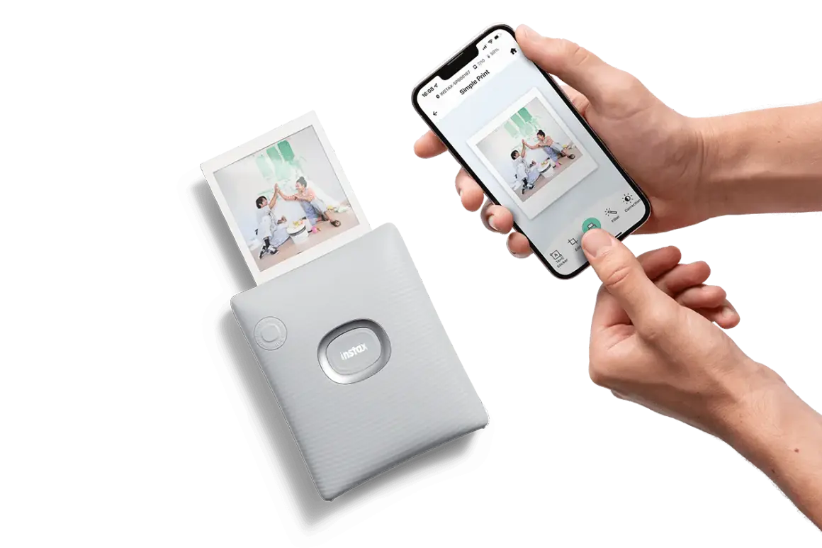 Ręce trzymają smartfon z aplikacją do drukowania zdjęć, obok drukarka instax wypuszcza gotowe zdjęcia jak z instax.