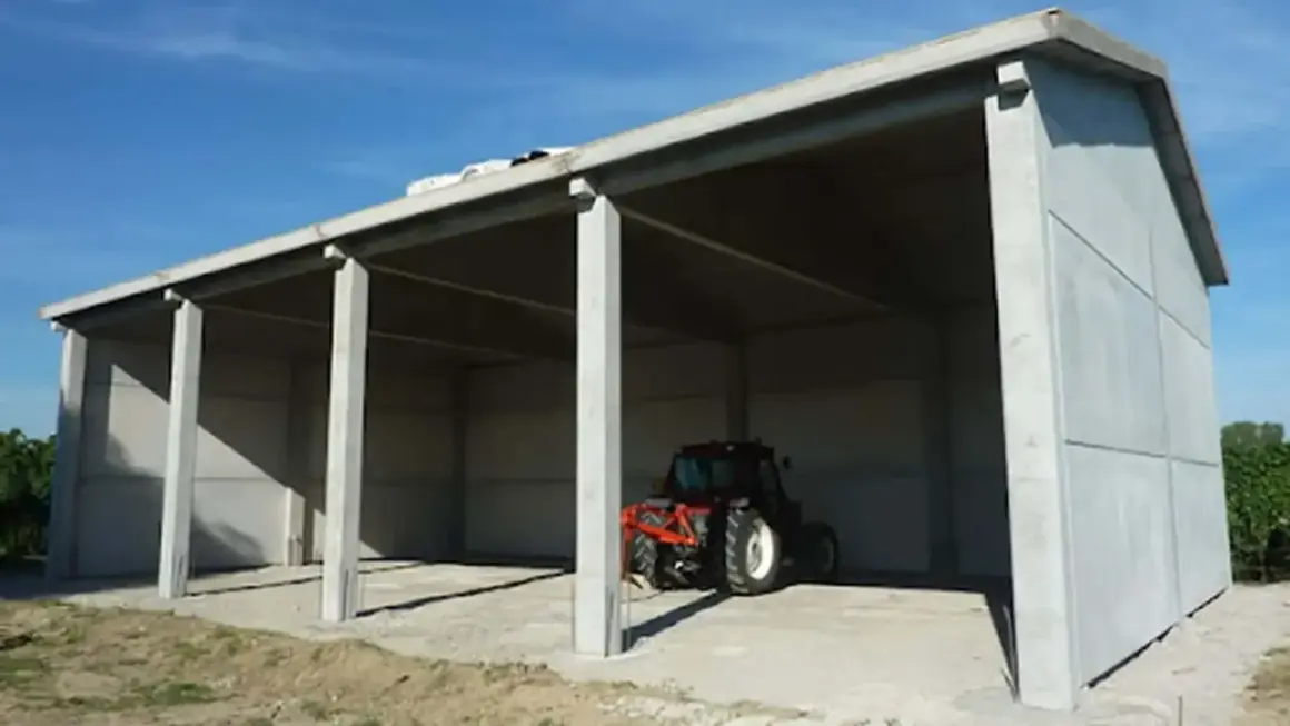 Capannone prefabbricato in cemento in zona agricola, ideale per chi vuole costruire in zona agricola senza essere imprenditore agricolo. Un trattore è parcheggiato all'interno.