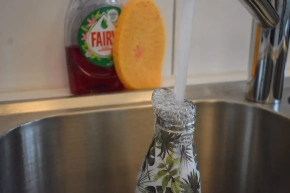 Eine Nalgene-Flasche mit Blattmuster wird in der Spüle unter fließendem Wasser gespült. Spülmittel und Schwamm sind im Hintergrund sichtbar.