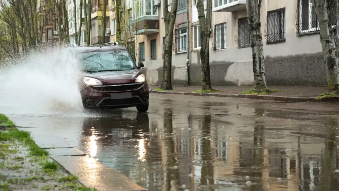 Czerwony samochód wpada w kałużę, tworząc fontannę wody. To przykład aquaplaningu, czyli utraty przyczepności na mokrej nawierzchni.