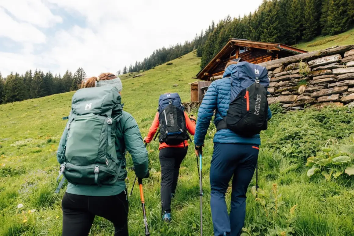 Tres excursionistas con mochilas y bastones suben por una ladera verde hacia una cabaña de madera. Saben qué llevar a la montaña.