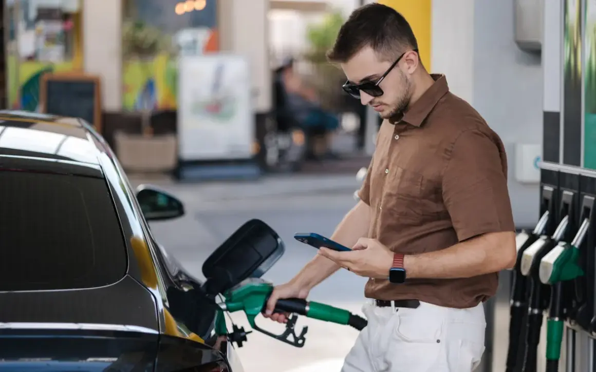 Un homme fait le plein de sa voiture à une **téléphone station essence**, consultant son smartphone.