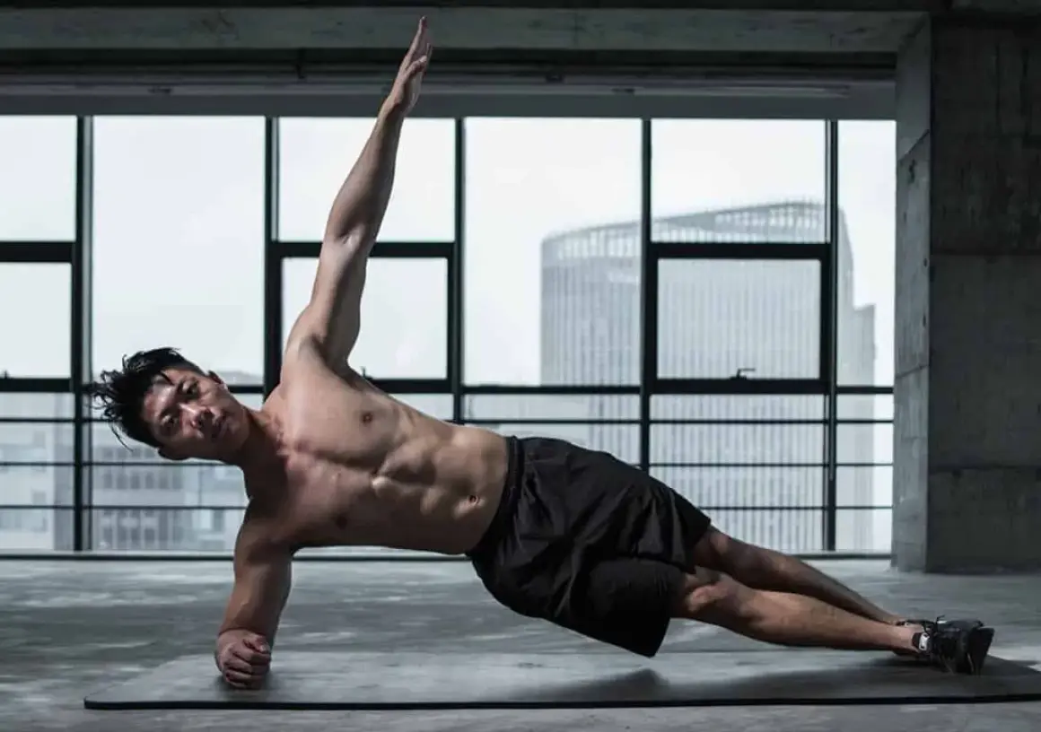 Un homme musclé réalise un exercice oblique, bras levé, dans une salle de sport avec vue sur la ville.