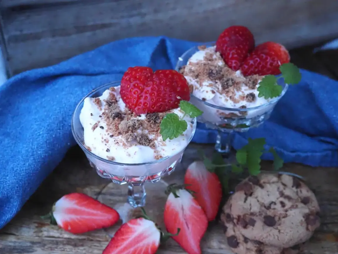 Två glas med krämig dessert toppad med jordgubbar och smulor. Bredvid ligger halverade jordgubbar och chokladkakor. En härlig sommardessert med jordgubbar och glass.
