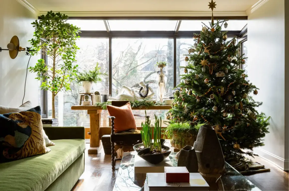 Cozy apartment Christmas decor features a decorated tree, green sofa, and a large window overlooking trees.