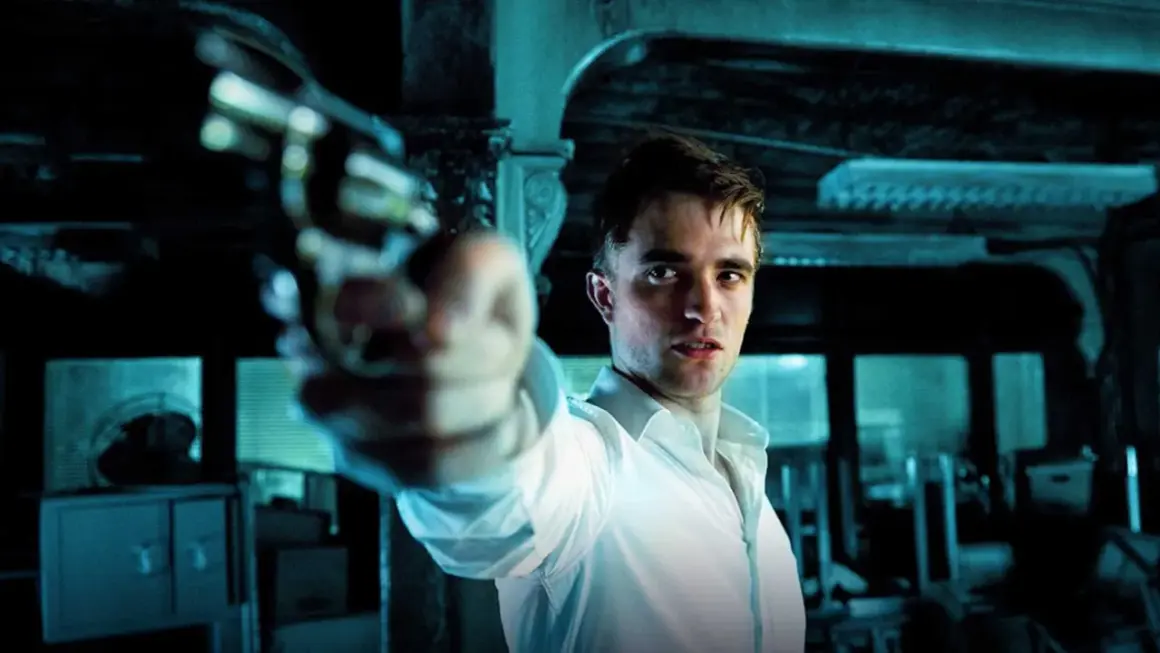 Robert Pattinson i en vit skjorta siktar med en pistol i en mörk, mystisk scen, typisk för hans filmer.