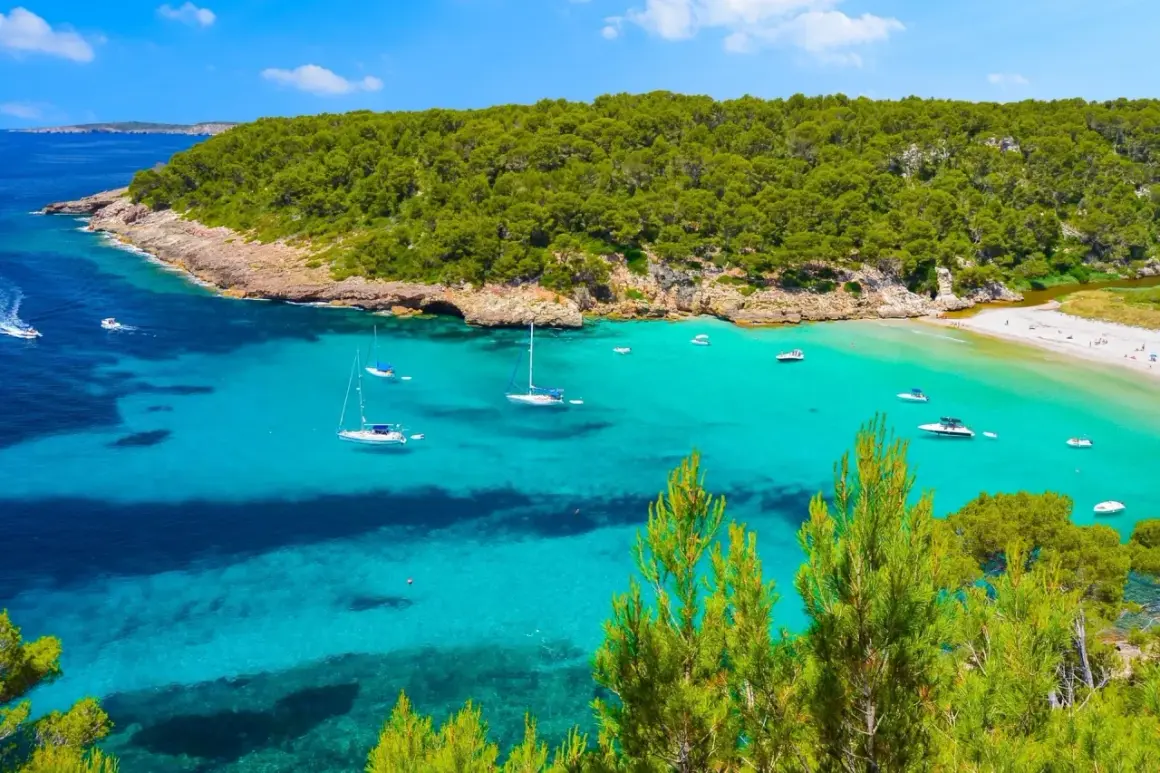 Bahía de aguas turquesas y cristalinas, con veleros y lanchas navegando. Al fondo, una playa de arena blanca y una frondosa vegetación que llega hasta la cala pudent.